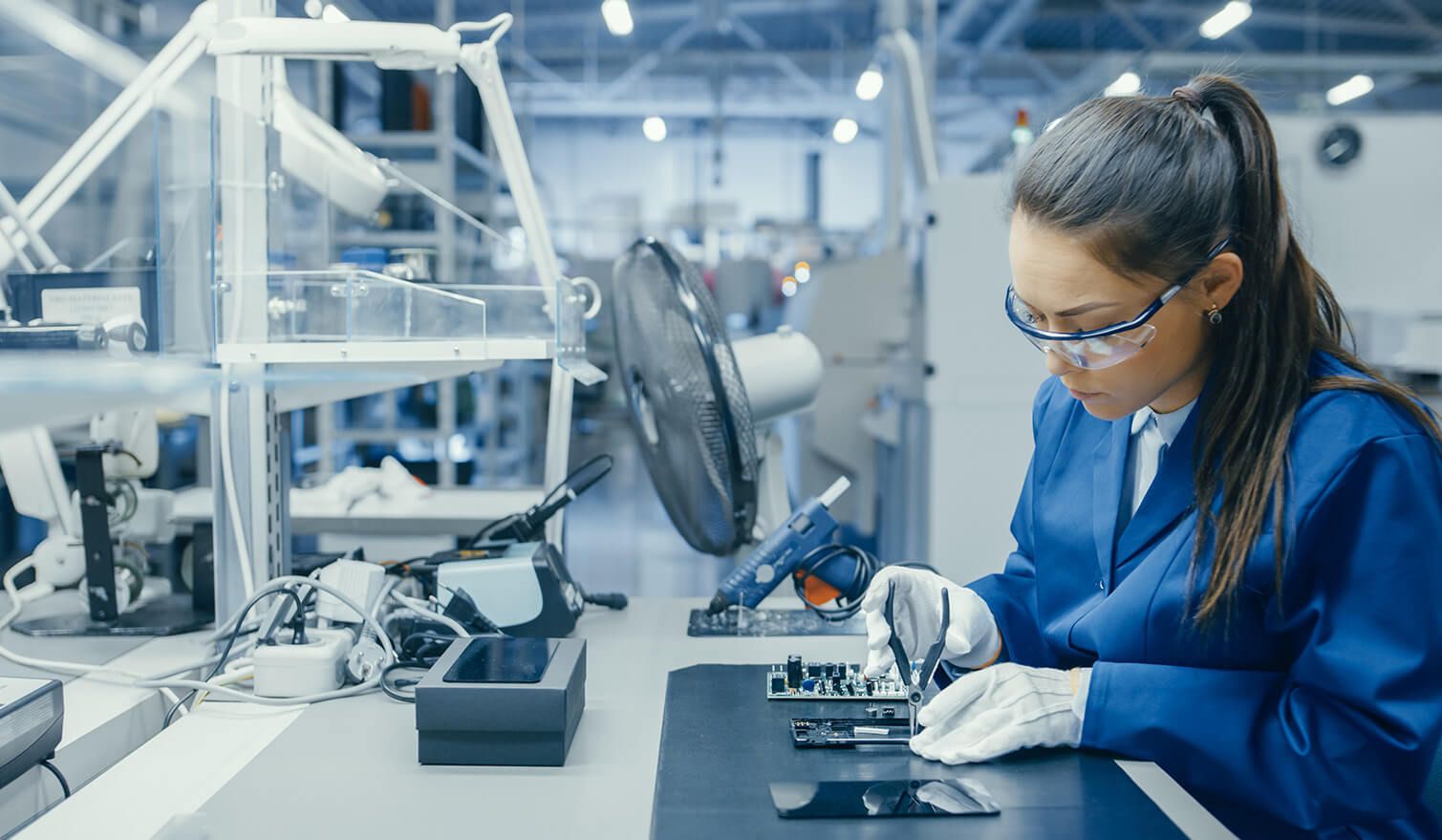 Kobieta w niebieskim stroju precyzyjnie obsługuje sprzęt elektroniczny w odpowiednio oświetlonym środowisku produkcyjnym